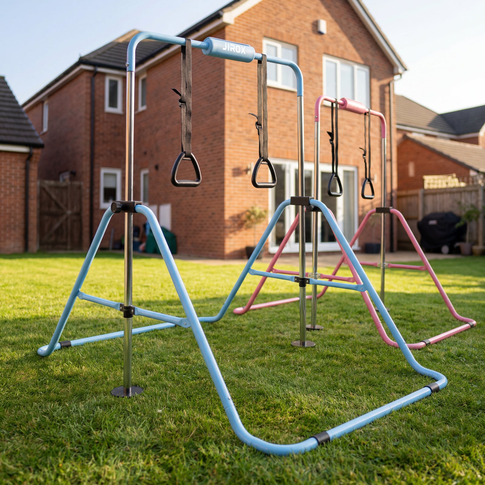 Freestanding kids' gym bar set with blue and pink frames and hanging gymnastic rings, set up on a garden lawn in front of a house