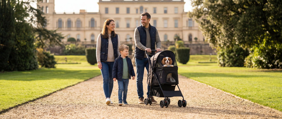 Family walking through a park with a young boy and a dog riding in a black pet stroller, with a large stately home in the background